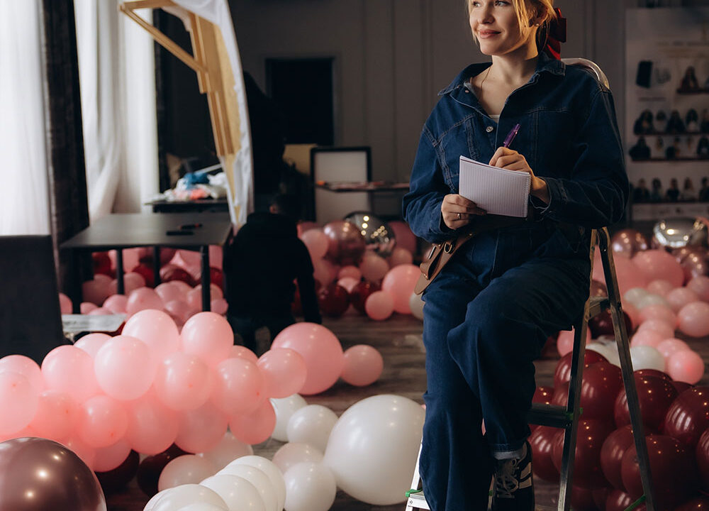 Event decor professional sitting on a step ladder with a notepad, surrounded by pink and burgundy balloons