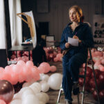 Event decor professional sitting on a step ladder with a notepad, surrounded by pink and burgundy balloons