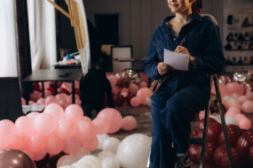 Event decor professional sitting on a step ladder with a notepad, surrounded by pink and burgundy balloons