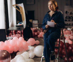 Event decor professional sitting on a step ladder with a notepad, surrounded by pink and burgundy balloons