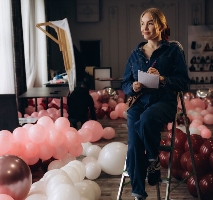Event decor professional sitting on a step ladder with a notepad, surrounded by pink and burgundy balloons