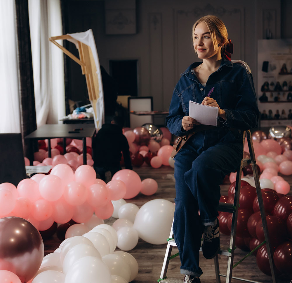 Event decor professional sitting on a step ladder with a notepad, surrounded by pink and burgundy balloons