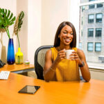 Small business owner smiling with a coffee mug at her office desk beside a monitor and a city window