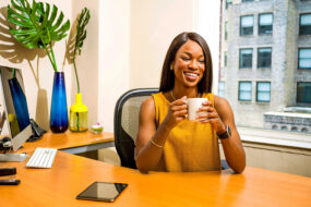 Small business owner smiling with a coffee mug at her office desk beside a monitor and a city window
