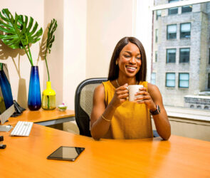 Small business owner smiling with a coffee mug at her office desk beside a monitor and a city window