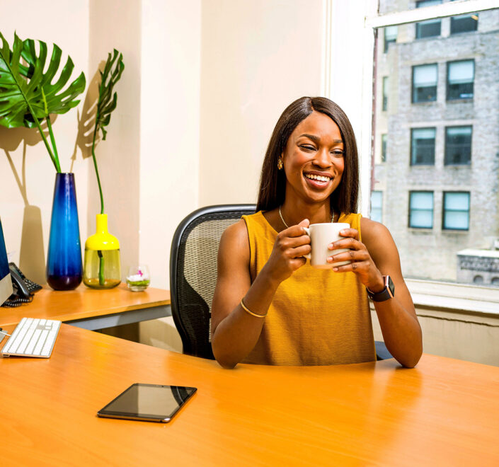 Small business owner smiling with a coffee mug at her office desk beside a monitor and a city window