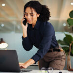Business consultant taking a phone call while standing over her laptop in a bright office