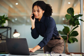 Business consultant taking a phone call while standing over her laptop in a bright office