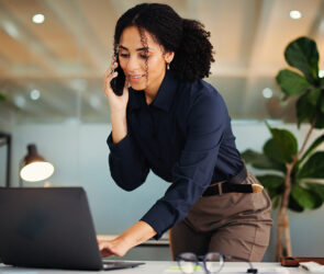 Business consultant taking a phone call while standing over her laptop in a bright office