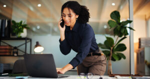 Business consultant taking a phone call while standing over her laptop in a bright office