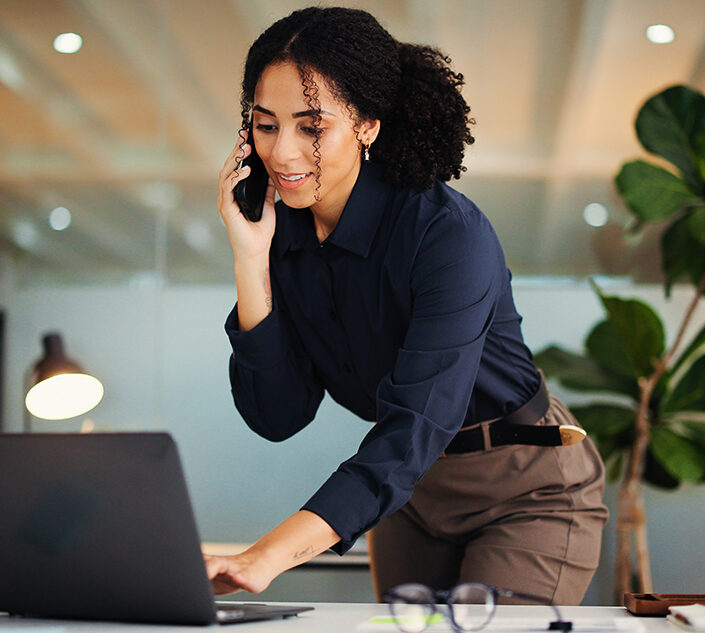 Business consultant taking a phone call while standing over her laptop in a bright office