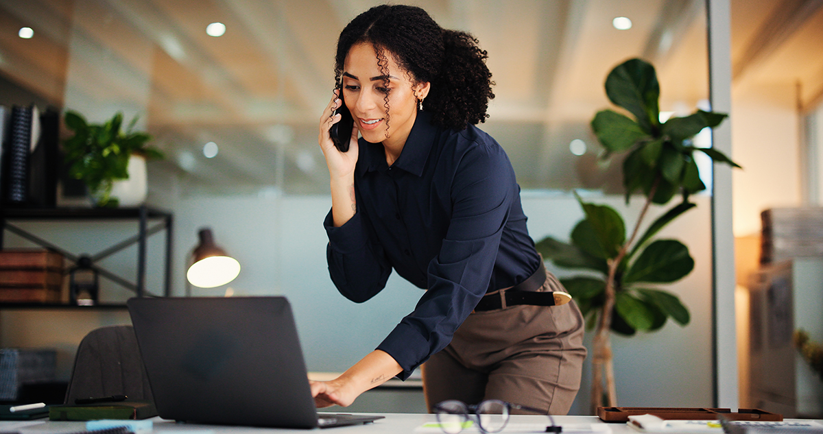 Business consultant taking a phone call while standing over her laptop in a bright office