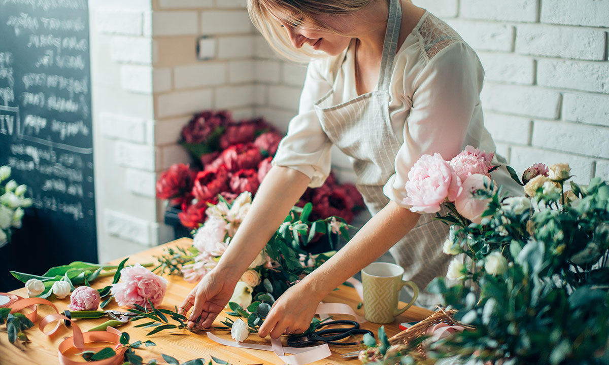 Florist arranging peonies and greenery on a wooden worktable with ribbon and scissors