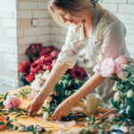 Florist arranging peonies and greenery on a wooden worktable with ribbon and scissors