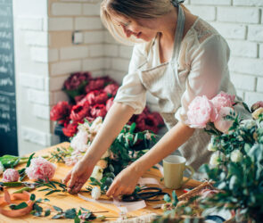 Florist arranging peonies and greenery on a wooden worktable with ribbon and scissors