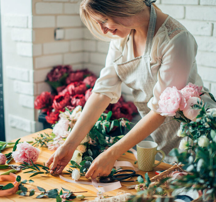 Florist arranging peonies and greenery on a wooden worktable with ribbon and scissors