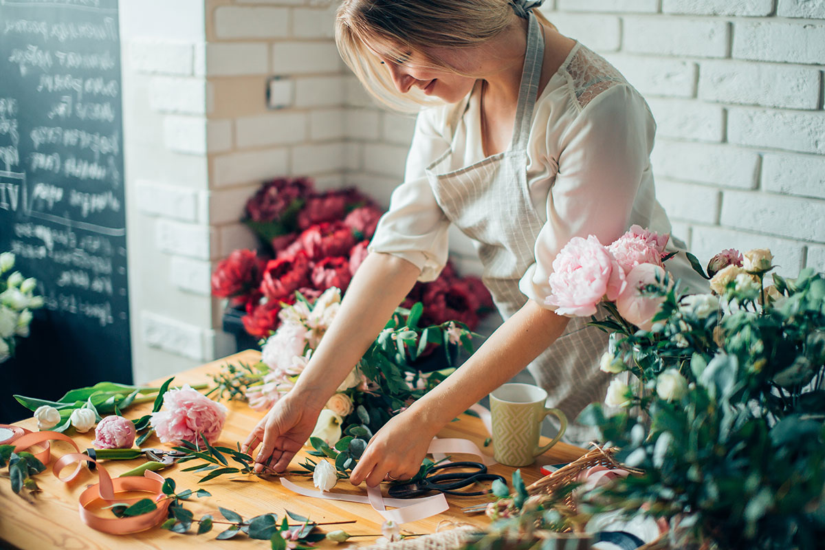 Florist arranging peonies and greenery on a wooden worktable with ribbon and scissors