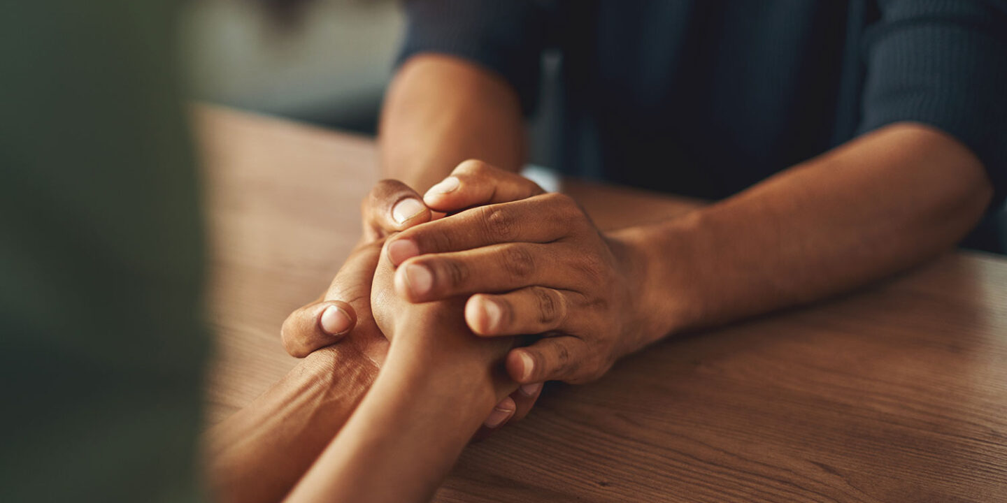 Two people holding hands across a wooden table in a moment of comfort and support