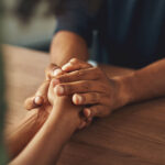 Two people holding hands across a wooden table in a moment of comfort and support