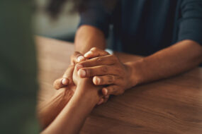 Two people holding hands across a wooden table in a moment of comfort and support