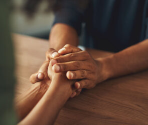 Two people holding hands across a wooden table in a moment of comfort and support