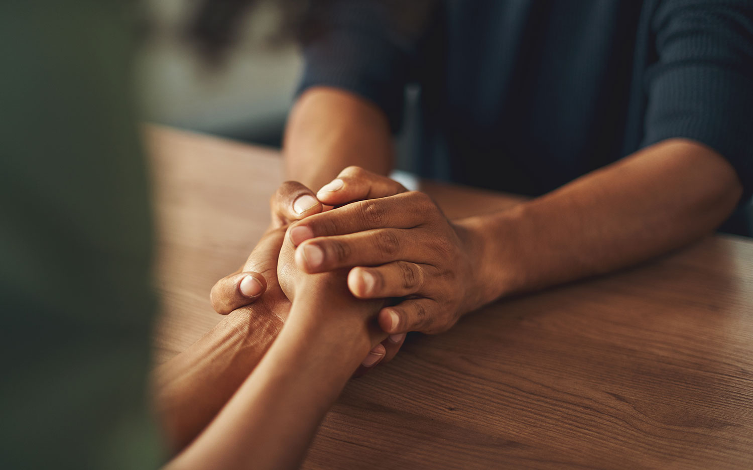 Two people holding hands across a wooden table in a moment of comfort and support