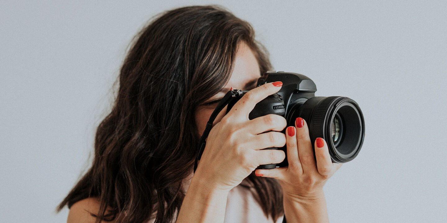 Portrait photographer with red nails looking through her camera against a plain light gray backdrop