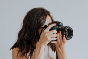Portrait photographer with red nails looking through her camera against a plain light gray backdrop