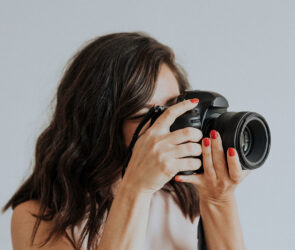 Portrait photographer with red nails looking through her camera against a plain light gray backdrop
