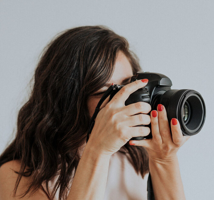 Portrait photographer with red nails looking through her camera against a plain light gray backdrop