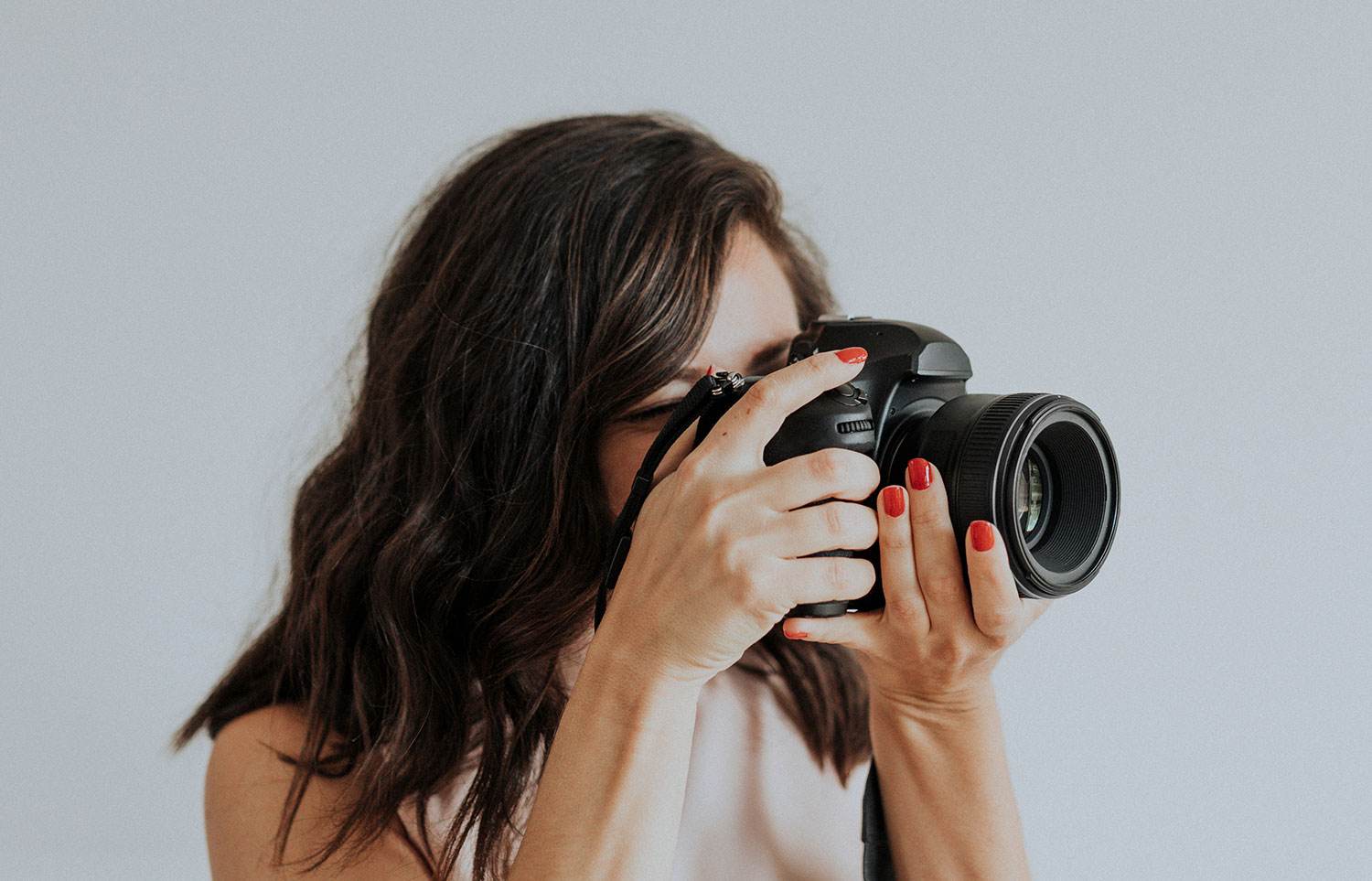 Portrait photographer with red nails looking through her camera against a plain light gray backdrop