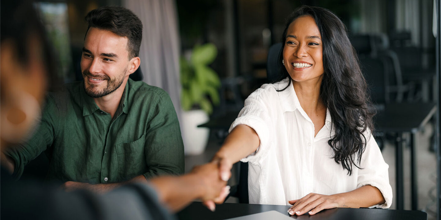 Smiling business owner shaking hands with a new client across a table, a colleague beside her and a closed laptop in front