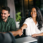 Smiling business owner shaking hands with a new client across a table, a colleague beside her and a closed laptop in front