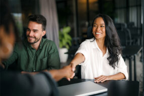 Smiling business owner shaking hands with a new client across a table, a colleague beside her and a closed laptop in front