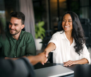 Smiling business owner shaking hands with a new client across a table, a colleague beside her and a closed laptop in front