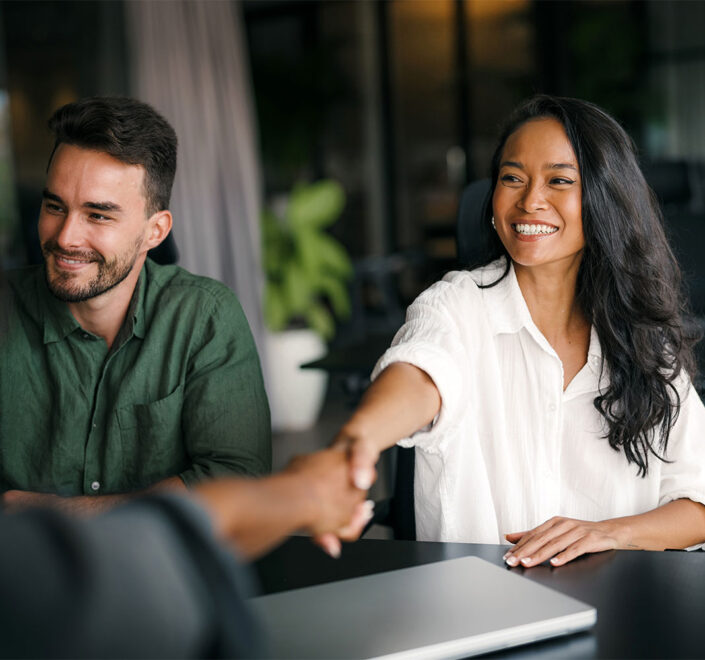Smiling business owner shaking hands with a new client across a table, a colleague beside her and a closed laptop in front