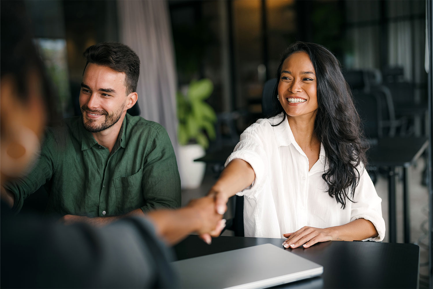 Smiling business owner shaking hands with a new client across a table, a colleague beside her and a closed laptop in front